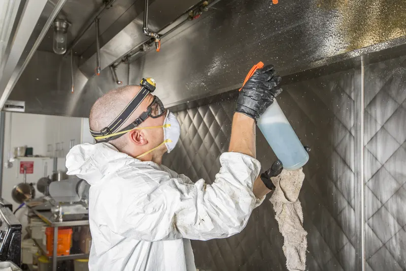 Technician spraying and wiping a hood interior with commercial cooking equipment behind